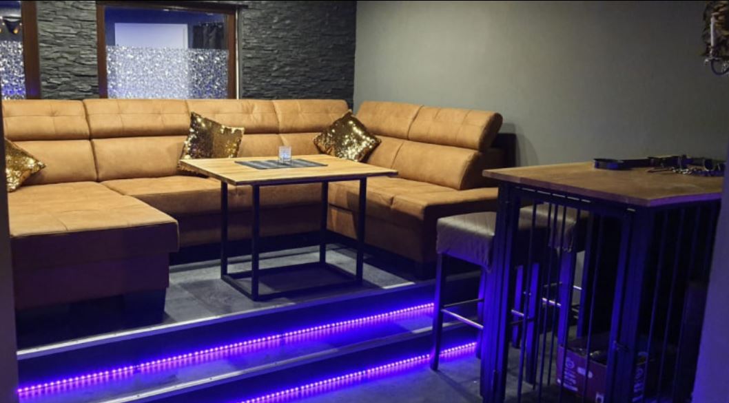 Lounge mit brauner Eckcouch, Holztisch und beleuchteter Stufe.